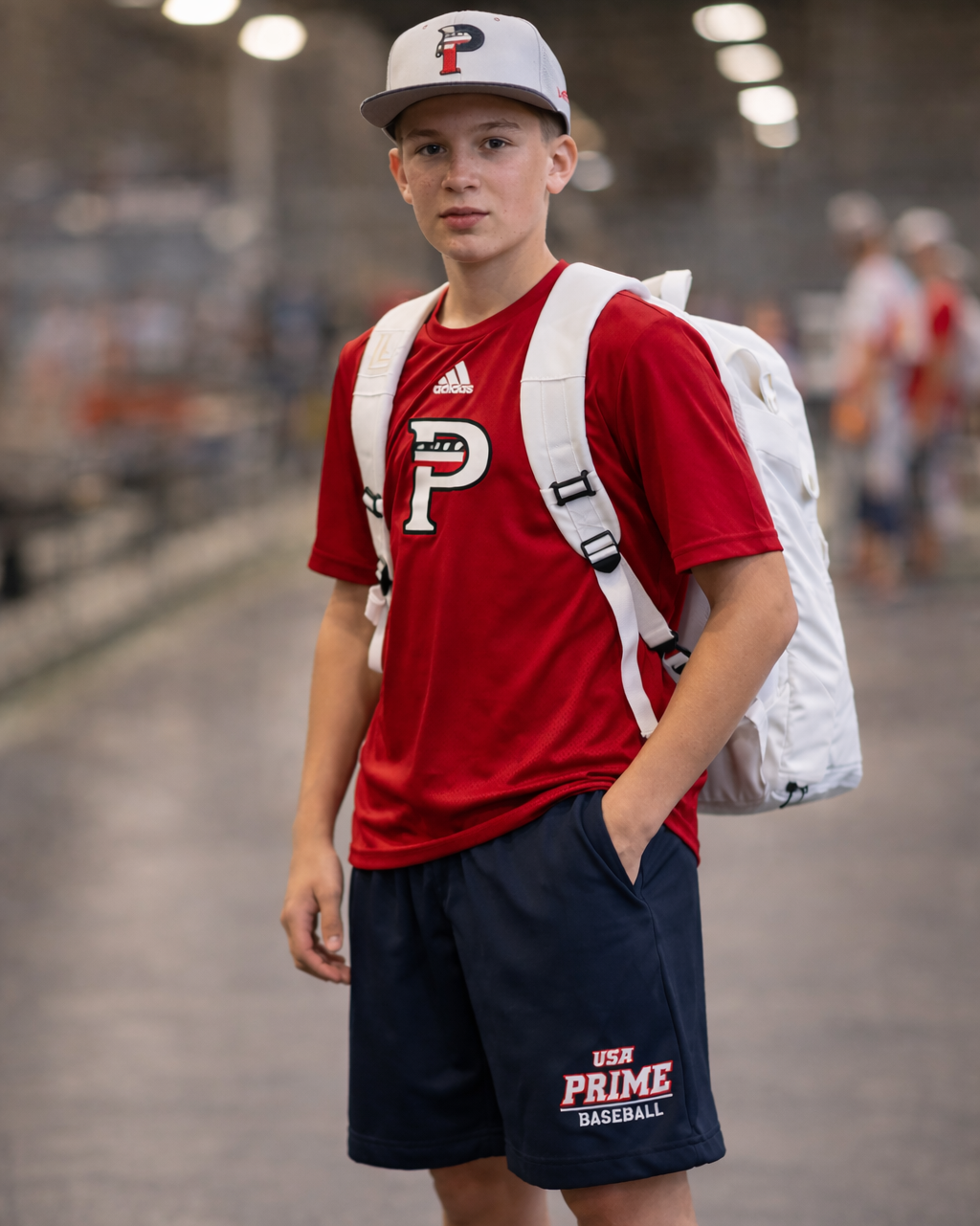 Young person wearing a red shirt and navy shorts with a white backpack, standing in an indoor setting.